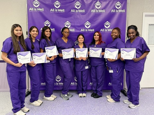 Eight graduates in purple scrubs holding certificates beside the All is Well banner