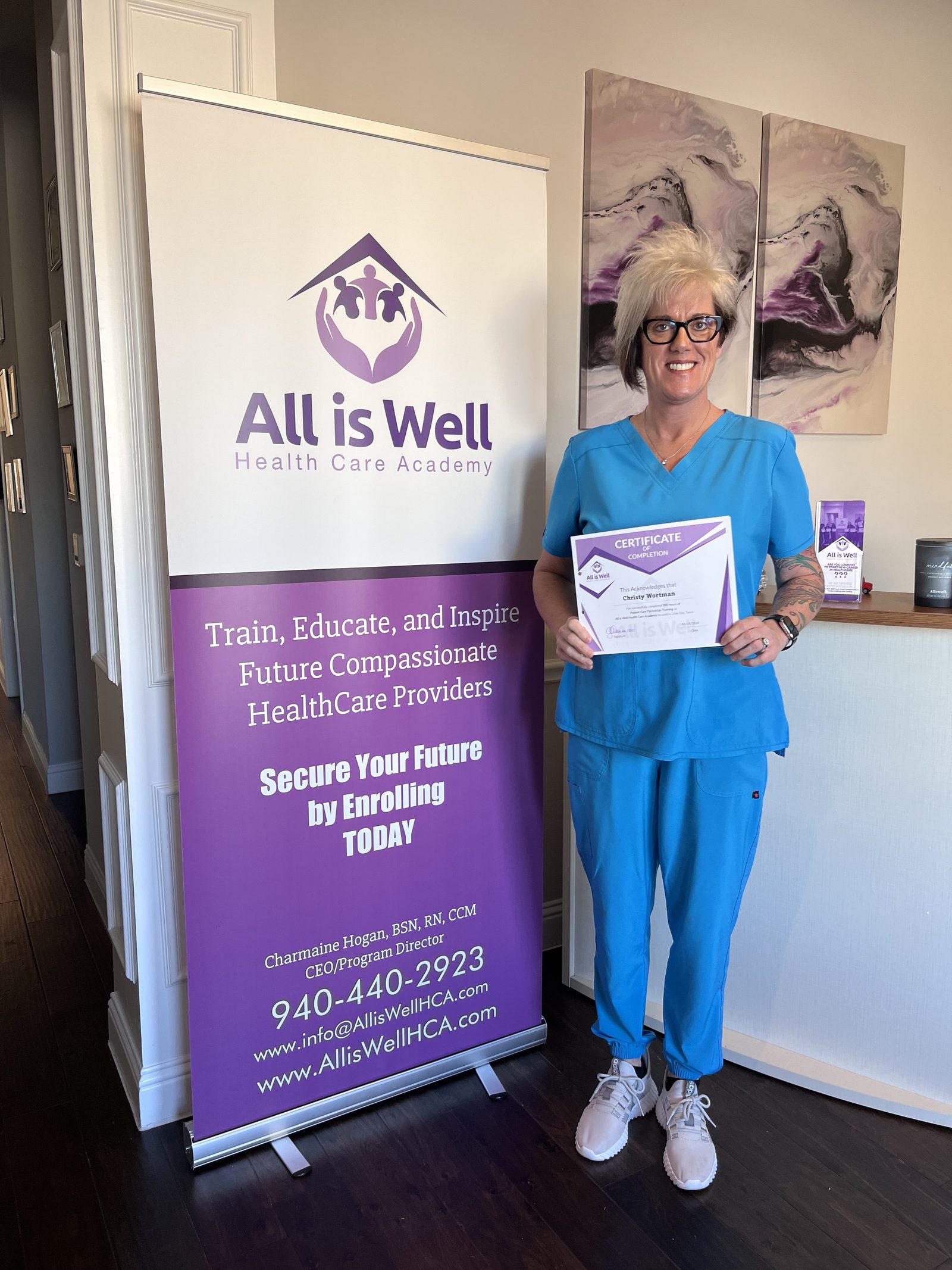 All is Well graduate holding a certificate of completion beside the academy banner